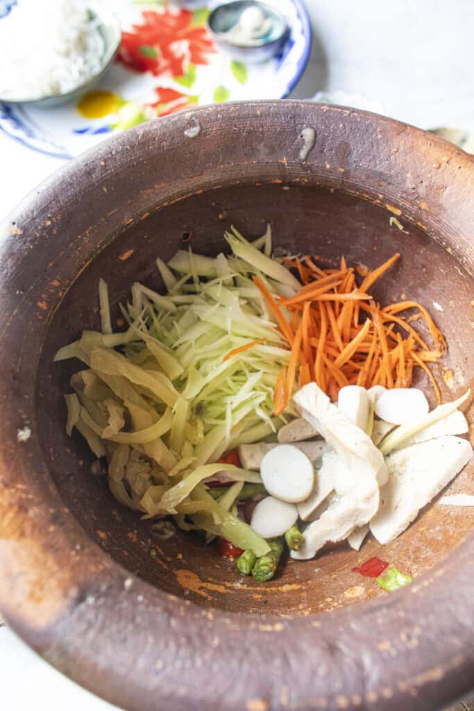 Shredded green papaya, carrot, and mustard greens added to Som Tum Mua in a clay mortar.