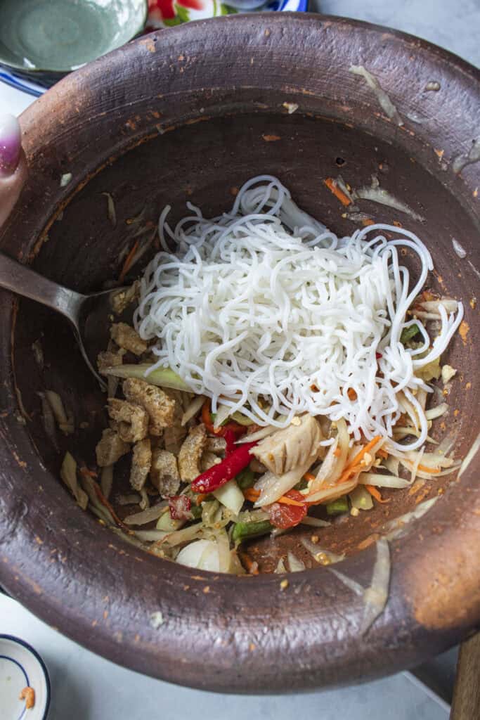Rice vermicelli noodles added to Som Tum Mua papaya salad in a clay mortar.