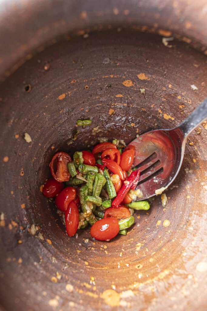 Pounding garlic, Thai chilies, beans and tomatoes in a clay mortar to start Som Tum Mua Thai papaya salad