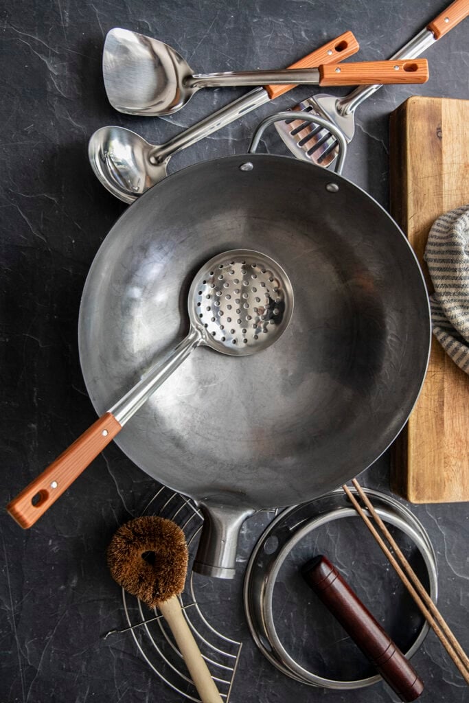 Carbon steel wok with wok utensils, chopsticks, wok ring, and scrubber on a dark countertop.